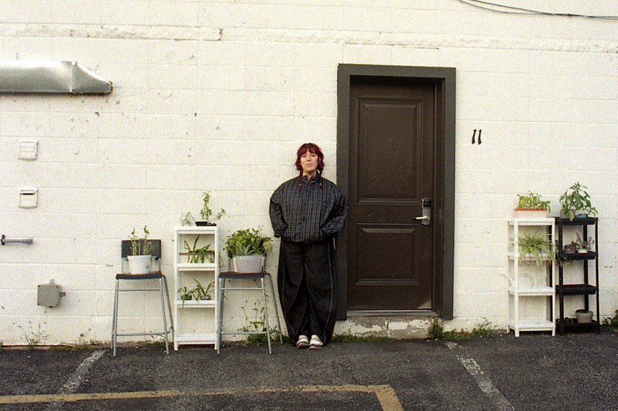 A colour photo shows the frontperson of the group Fanclubwallet standing outside of a motel room againt a white brick wall and to the left of a brown door numbered 11. Chairs and stands containing potted plants are on each side of them. 