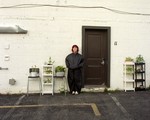 A colour photo shows the frontperson of the group Fanclubwallet standing outside of a motel room againt a white brick wall and to the left of a brown door numbered 11. Chairs and stands containing potted plants are on each side of them. 
