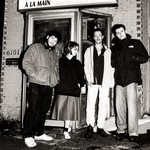 Four members of Fionavair stand outside a small brick building, dressed in casual cool‑weather clothing. They pose in front of a glass door beneath a sign reading “Lave Auto à la Main,” creating a candid, black‑and‑white street‑style band portrait.