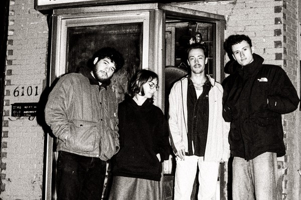 Four members of Fionavair stand outside a small brick building, dressed in casual cool‑weather clothing. They pose in front of a glass door beneath a sign reading “Lave Auto à la Main,” creating a candid, black‑and‑white street‑style band portrait.