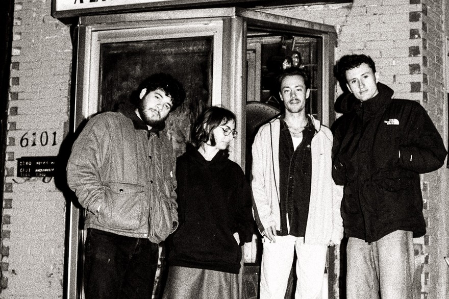 Four members of Fionavair stand outside a small brick building, dressed in casual cool‑weather clothing. They pose in front of a glass door beneath a sign reading “Lave Auto à la Main,” creating a candid, black‑and‑white street‑style band portrait.