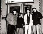 Four members of Fionavair stand outside a small brick building, dressed in casual cool‑weather clothing. They pose in front of a glass door beneath a sign reading “Lave Auto à la Main,” creating a candid, black‑and‑white street‑style band portrait.