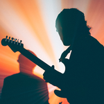 Two members of Forest Ray are shown playing drums and guitar backlit in front of vibrant red a yellow lights, showing only their silhouettes.