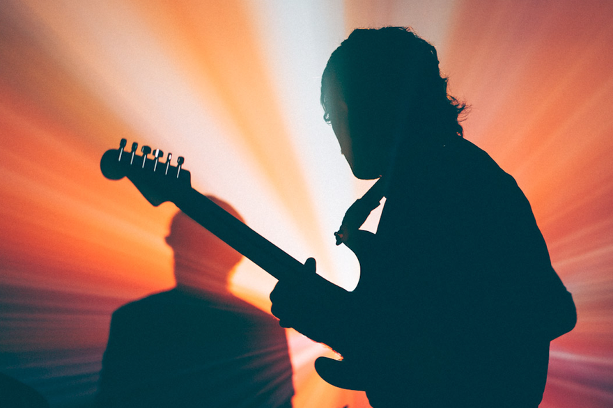 Two members of Forest Ray are shown playing drums and guitar backlit in front of vibrant red a yellow lights, showing only their silhouettes.