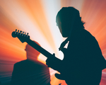 Two members of Forest Ray are shown playing drums and guitar backlit in front of vibrant red a yellow lights, showing only their silhouettes.