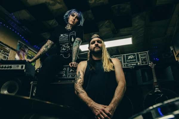 A colour photo taken from below shows the duo FRANKLIN sitting in what looks like a jam space filled with amps and guitars. 
