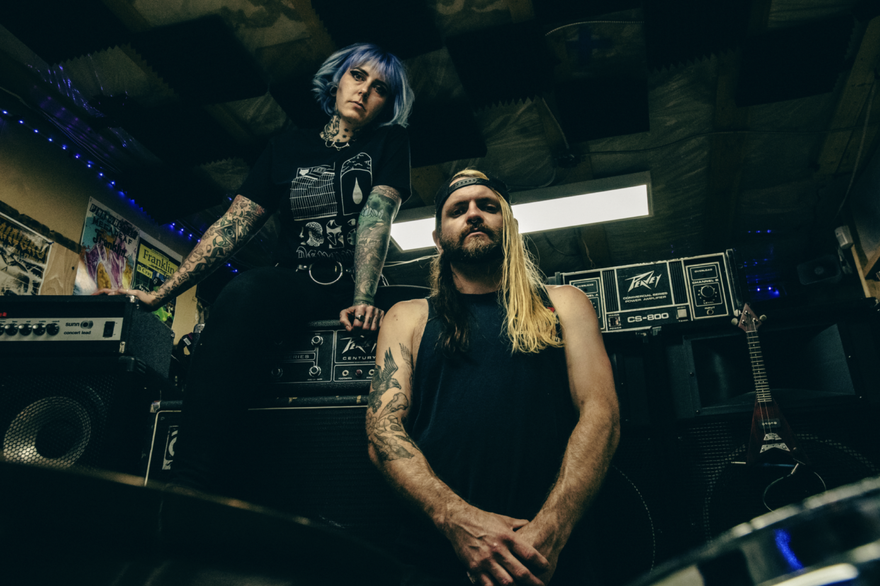 A colour photo taken from below shows the duo FRANKLIN sitting in what looks like a jam space filled with amps and guitars. 