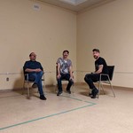 Three members of the band Fulfilment sit on black chairs in a empty beige room and look at the camera.