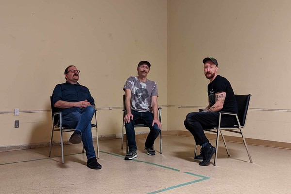 Three members of the band Fulfilment sit on black chairs in a empty beige room and look at the camera.