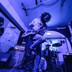 A photo bathed in purple light shows four-piece rock band performing in a small venue with two guitars, a bass and drums. 