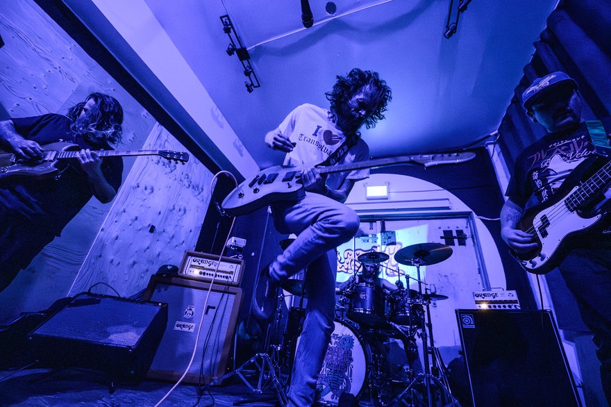 A photo bathed in purple light shows four-piece rock band performing in a small venue with two guitars, a bass and drums. 