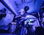 A photo bathed in purple light shows four-piece rock band performing in a small venue with two guitars, a bass and drums. 