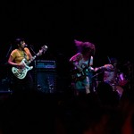 A colour photo taken from the crowd shows three-piece band Gadfly performing onstage with two guitars, drums and amps in the background. 