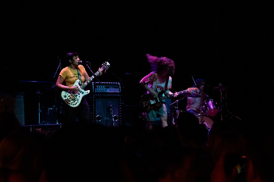 A colour photo taken from the crowd shows three-piece band Gadfly performing onstage with two guitars, drums and amps in the background. 