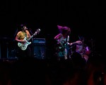 A colour photo taken from the crowd shows three-piece band Gadfly performing onstage with two guitars, drums and amps in the background. 