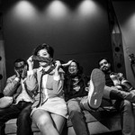 A black and white photo shows the four members of the band Ginger Beef sitting on a couch with guitars on each side. One member second from the left holds a tie in front of their face covering their nose. 