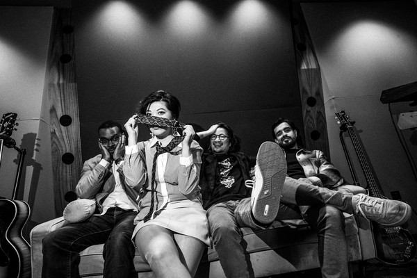 A black and white photo shows the four members of the band Ginger Beef sitting on a couch with guitars on each side. One member second from the left holds a tie in front of their face covering their nose. 