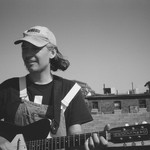 A close-up black and white photo shows artist Gus Englehorn playing guitar on a rooftop with other buildings seen in the background. 