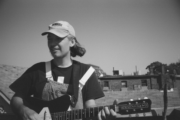A close-up black and white photo shows artist Gus Englehorn playing guitar on a rooftop with other buildings seen in the background. 
