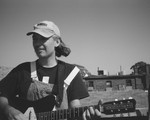 A close-up black and white photo shows artist Gus Englehorn playing guitar on a rooftop with other buildings seen in the background. 