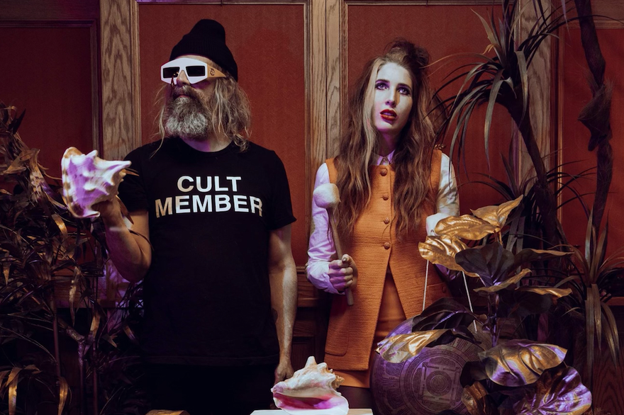 A colour photo shows duo Hair Control in a decorated room with gold plants and a white pillar in the centre. The member on the left holds a conch shell and wears a black t-shirts that read cult member. The member on the right wears an orange vest and is seen holding a marshmellow on a stick.