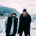 A slightly overexposed colour photo shows the duo Halfcut & Cole The God standing in a snowy field with mountains and pine trees in the background. 