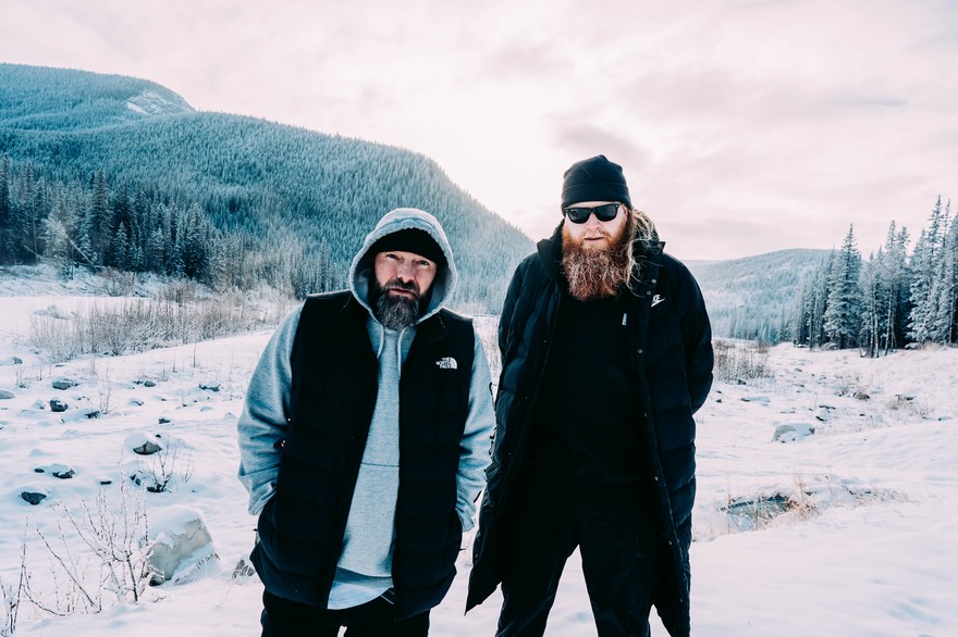 A slightly overexposed colour photo shows the duo Halfcut & Cole The God standing in a snowy field with mountains and pine trees in the background. 