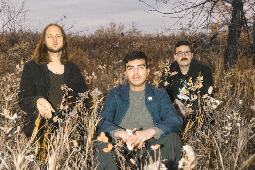 The three members of the group Heavydive are shown sitting in a field during autumn with a grey cloudy sky behind them and a tree to the right.
