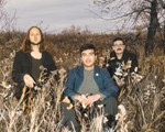 The three members of the group Heavydive are shown sitting in a field during autumn with a grey cloudy sky behind them and a tree to the right.