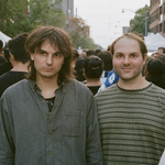A colour photo of duo Heaven For Real shows them standing in the middle of a busy street looking the towards the camera while the rest of the crowd walks by them in the opposite direction. 