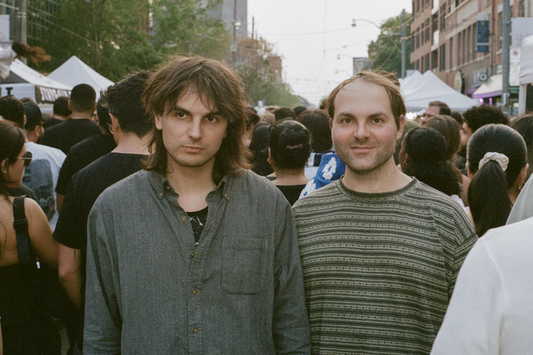 A colour photo of duo Heaven For Real shows them standing in the middle of a busy street looking the towards the camera while the rest of the crowd walks by them in the opposite direction. 