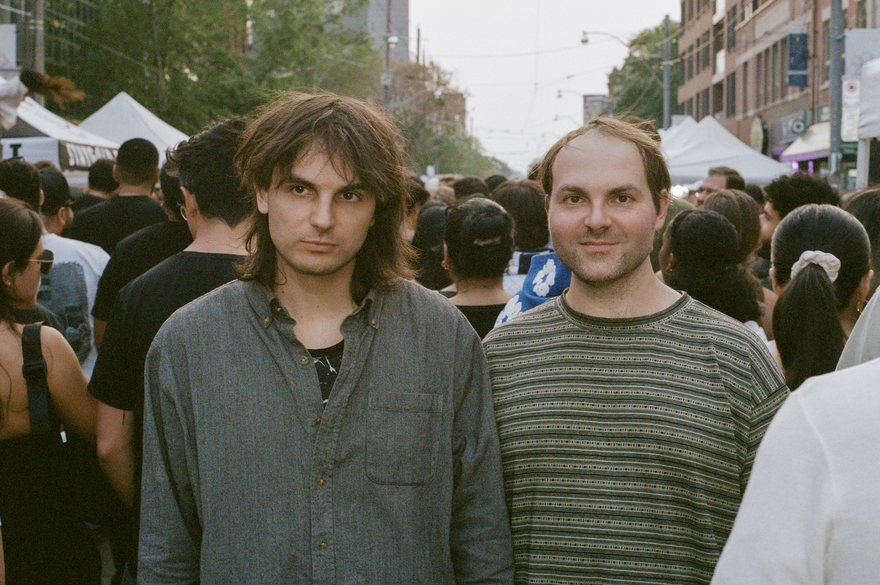 A colour photo of duo Heaven For Real shows them standing in the middle of a busy street looking the towards the camera while the rest of the crowd walks by them in the opposite direction. 