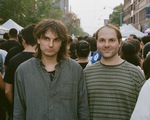 A colour photo of duo Heaven For Real shows them standing in the middle of a busy street looking the towards the camera while the rest of the crowd walks by them in the opposite direction. 