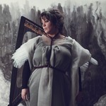 A colour photo shows artist Hermitess in a grey dress standing in front of a harp with a grey abstract painted wall shown behind them.