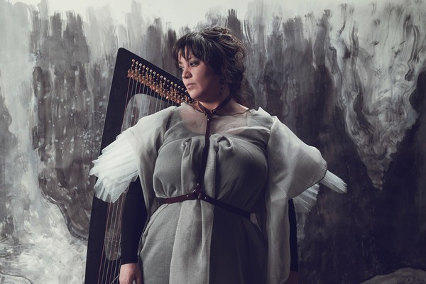 A colour photo shows artist Hermitess in a grey dress standing in front of a harp with a grey abstract painted wall shown behind them.