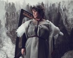A colour photo shows artist Hermitess in a grey dress standing in front of a harp with a grey abstract painted wall shown behind them.