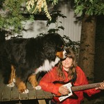 A colour photo shows artist Helene Barbier sitting on the right side of a picnic table with a bright red coat playing guitar. A bernese dog stands on the table with its head blocking their view. 