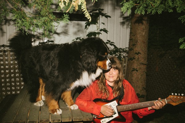 A colour photo shows artist Helene Barbier sitting on the right side of a picnic table with a bright red coat playing guitar. A bernese dog stands on the table with its head blocking their view. 