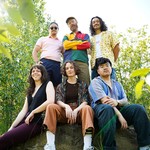 A colour photo shows six-piece band Hip Hip Thursday standing outside with leafy trees behind them. Three members sit on a rock in the foreground, while three members stand behind them. 