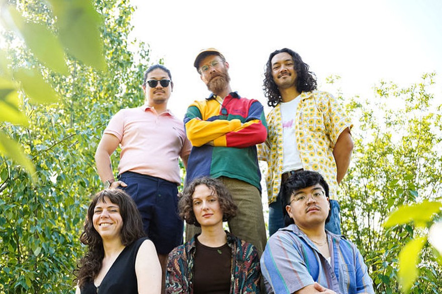 A colour photo shows six-piece band Hip Hip Thursday standing outside with leafy trees behind them. Three members sit on a rock in the foreground, while three members stand behind them. 