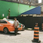 A colour photo shows the solo member of project His His standing in a parking lot leaning on an orange car. A bright green brick wall and other buildings are seen behind them. Three pylons are shown on the right hand side. 