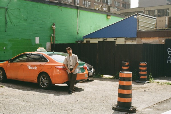 A colour photo shows the solo member of project His His standing in a parking lot leaning on an orange car. A bright green brick wall and other buildings are seen behind them. Three pylons are shown on the right hand side. 