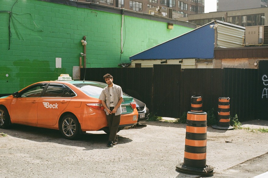 A colour photo shows the solo member of project His His standing in a parking lot leaning on an orange car. A bright green brick wall and other buildings are seen behind them. Three pylons are shown on the right hand side. 