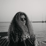 A black and white photo shows artist Ice Tha One seated on a metal grate with a body of water behind them and several sailboats in the distance. 