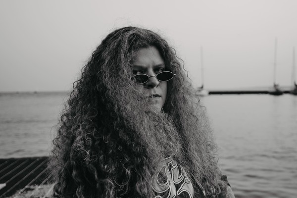 A black and white photo shows artist Ice Tha One seated on a metal grate with a body of water behind them and several sailboats in the distance. 