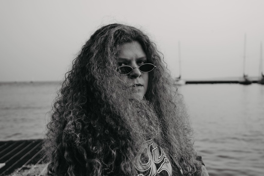 A black and white photo shows artist Ice Tha One seated on a metal grate with a body of water behind them and several sailboats in the distance. 