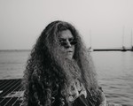 A black and white photo shows artist Ice Tha One seated on a metal grate with a body of water behind them and several sailboats in the distance. 