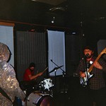 A grainy candid photo shows three-piece band Invisible Hand performing in a dark room with curtains drawn over the windows. 