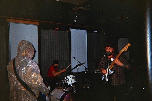 A grainy candid photo shows three-piece band Invisible Hand performing in a dark room with curtains drawn over the windows. 