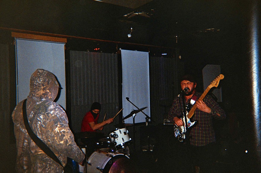 A grainy candid photo shows three-piece band Invisible Hand performing in a dark room with curtains drawn over the windows. 